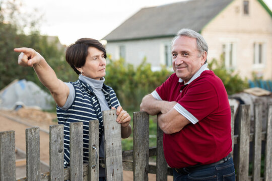 Old man and woman standing and talking near wooden fence in their garden