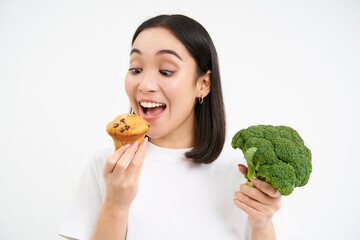 Close up of asian woman holding broccoli but eating tasty cupcake, white studio background