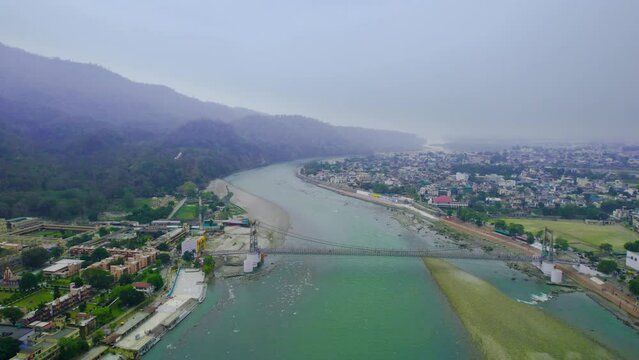 aerial drone shot flying towards janaki setu suspension bridge over blue water of river ganga stretching off into the distance between the himalaya mountain peaks with temples dotting the banks