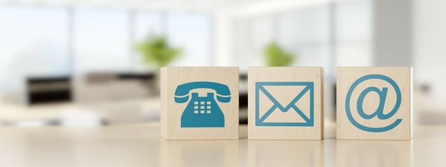 Wooden cube or box telephone, envelope letter and e-mail symbols in a row on wooden table with an empty blurred office in the background, contact us symbols or banner with copy space