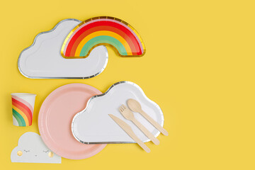 Cute paper party plates in the shape of rainbow and clouds with cups for themed kids party. Birthday party decorations on yellow background. Set of holiday disposable tableware for party or picnic.