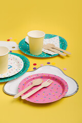 Cute paper party plates and cups for themed kids party on yellow background. Birthday party decorations and props in bright colors. Set of holiday disposable tableware for party or picnic.