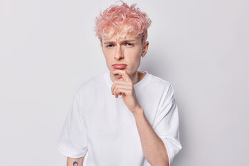 Negative facial expressions concept. Displeased pink haired European man keeps hand on chin frowns face feels discontent dressed in casual t shirt isolated over white background. Studio shot