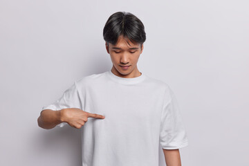 Horizontal shot of young Chinese man points at blank copy space of casual t shirt shows space for advertising content isolated over white background. Clothing fashion design promotion concept