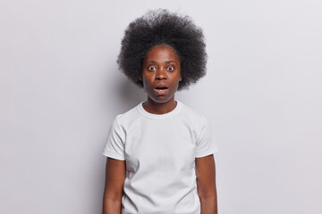 Omg concept. Curly haired woman with curly bushy hair sees something astonishing hears something unexpected dressed in casual t shirt isolated over white background being afraid of scarying news