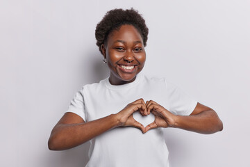 Lovely dark skined woman expresses sympathy and admiration makes heart gesture smiles toothily has romantic mood dressed in t shirt isolated over white background. Be my valentine. I love you sign