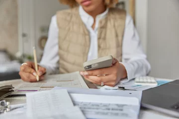 Fototapeten Cropped shot of unrecognizable woman holds mobile phone checks documents utility bills writes down information in notepad poses at table indoors surrounded by paper invoices counts home expenses © Wayhome Studio