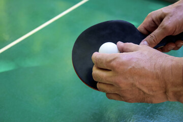 Man’s hands holds black ping pong racket and white tennis ball over green tennis table. Close-up....