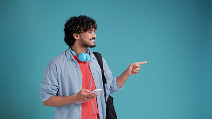 young latin spanish man with headphones and smartphone pointing with his finger to the side to the space for text, on a blue studio background