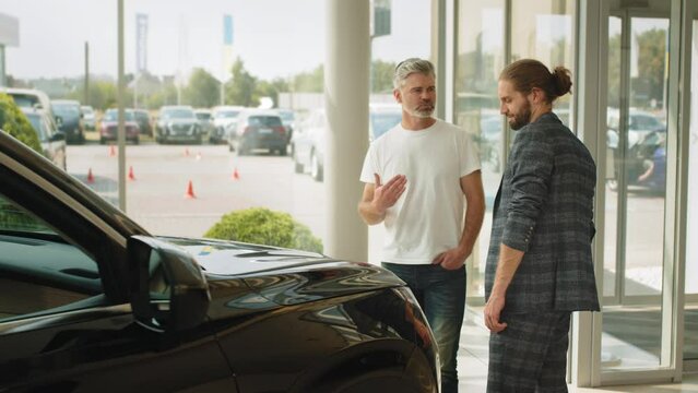 Two men elderly father and adult son in a showroom choose a new car. Young caucasian cars salesman show the best variant to customer in dealership. Handsome business man came to get new auto.