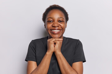 Portrait of cheerful dark skinned woman with short hair keeps hands under chin smiles toothily dressed in casual black t shirt expesses sincere positive emotions isolated over white background