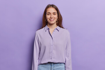 Portrait of dark haired woman smiles pleasantly keeps arms down feels happy to have first day at work dressed in shirt and jeans isolated over purple background. People and positive emotions concept
