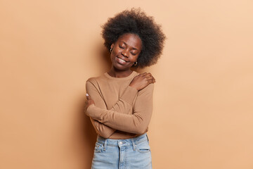 Curly haired woman recalls romantic event embraces herself with both arms tilts head smiles gently stands satisfied wears casual jumper jeans isolated on brown background expresses self love and care