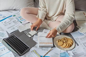 Cropped faceless woman manages family budget calculates bills household expenses surrounded by papers sits crossed legs on comfortable bed uses laptop computer makes online payment analyzes profit.
