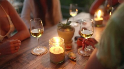 Diverse female friends drink wine in rooftop sea view restaurant at sunset. Multiracial women talking and smiling with candles at table in a tropical cafe at dusk. Young multiethnic women dining. - Powered by Adobe