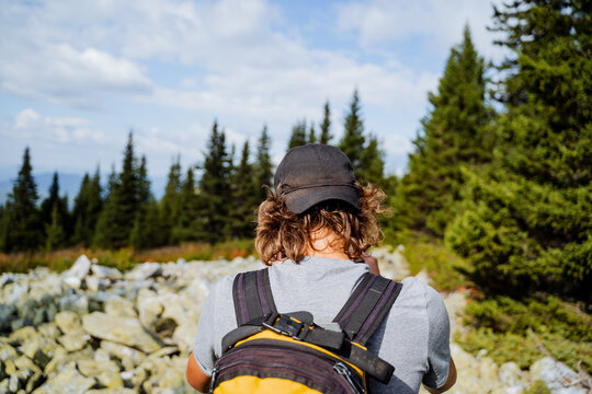 Rear view of a tourist man in the mountains, a black cap on his head, curly hair, a baseball cap, the back of a guy with a backpack, a taiga forest, one in the mountains.