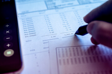 hand holding pen point at financial chart with accounting data paper and calculator in smartphone