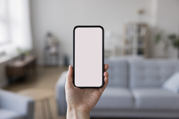 Close up of smartphone in female hand with blank empty screen. Woman holding mobile phone with copy space on display for online smart application interface. Cozy living room interior in background