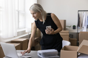 Busy senior entrepreneur woman using gadgets for processing Internet store order, sending product to client, packing cardboard box at home storage place, holding smartphone, typing on laptop