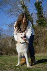Caucasian woman walks in field with white dog of mixed breed. Female owner has fun with pet outside in clearing near forest in spring. Girl with wavy hair strokes and hugs half breed Swiss Shepherd.