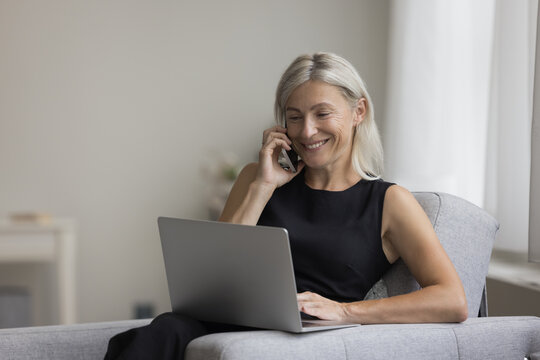 Happy positive blonde mature professional woman talking on mobile phone at laptop at home, enjoying online wireless communication, connection, resting in armchair, smiling, laughing