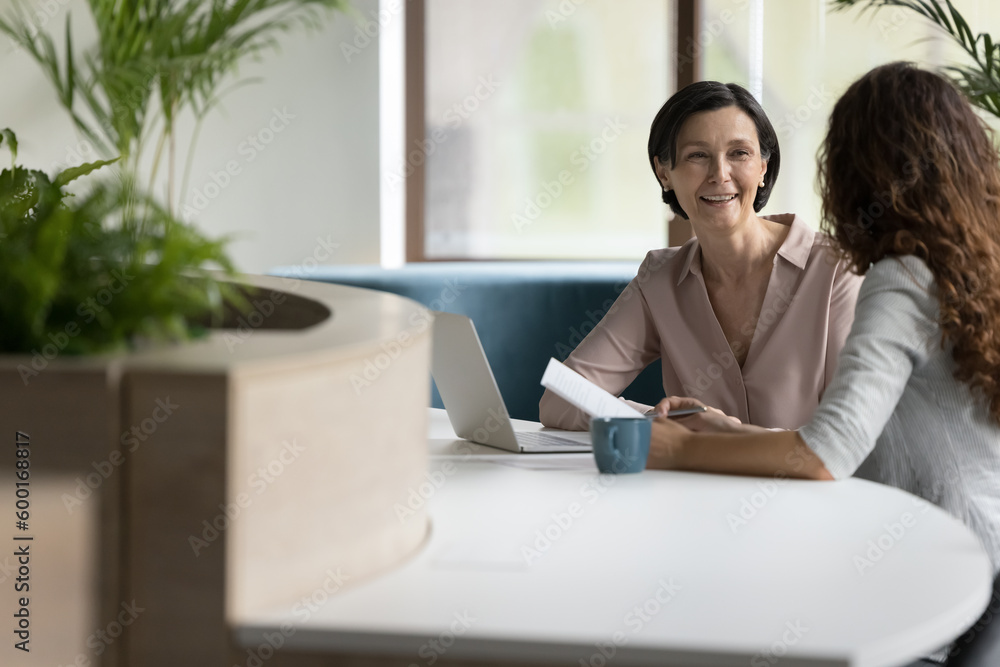 Wall mural Cheerful businesswoman talk to colleague, company client, met in office sit at desk with laptop, discuss paperwork, contract details, sell services to customer take part in formal meeting, do business