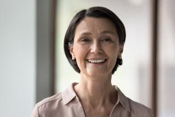 Close up head shot beautiful brunette mature businesswoman look at camera pose indoors, having wide...