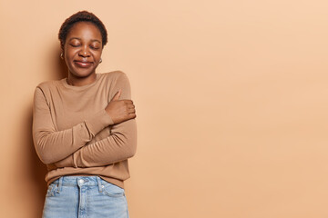 Black young girl embraces herself feels self pride stands with closed eyes smiles pleasantly dressed in casual jumper and jeans isolated over brown background blank space for your promotion.
