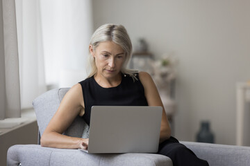 Focused mature customer woman using online service, app on laptop, shopping on Internet at home, relaxing in armchair, typing. Serious freelancer lady working on online project