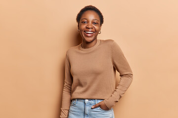 Studio shot of happy teenage girl with short hair keeps hand in pocket smiles broadly feels carefree dressed in casual jumper and jeans isolated over brown background. People and positive emotions
