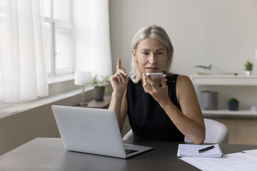 Serious busy mature freelancer business woman having telephone call on speaker, recording audio message on smartphone at laptop. Entrepreneur using digital gadgets for work communications at home