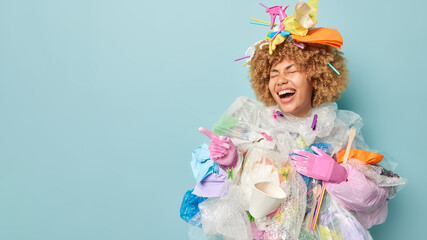 Overjoyed woman covered with plastic rubbish wears protective gloves points finger aside on blank space shows something funny collects garbage to clean out our planet stands against blue background