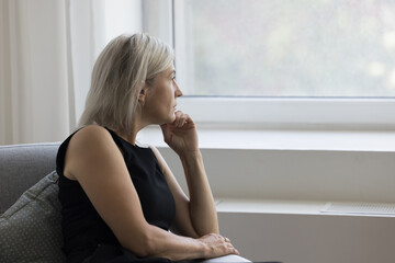 Thoughtful depressed mature woman sitting on couch at home, looking at window away in deep...