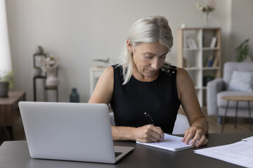 Serious mature retired woman studying at laptop at home, writing notes in notebook, watching online training webinar, professional course, listening to audio lecture