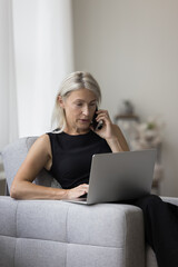Confident mature older business woman working from home, talking on cellphone, typing on laptop computer in armchair, looking at display, using Internet wireless communication for job
