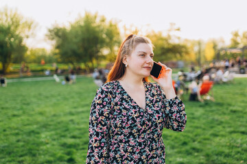 A portrait of a smiling beautiful woman talking on phone on nature background. Happy woman in dress is using a smartphone in park outdoors, summer time. Traveler