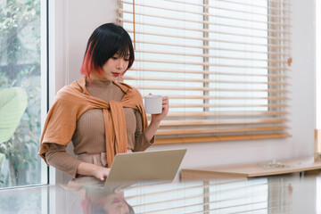 A beautiful Asian freelancer businesswoman is using a digital laptop near a window in her home office.