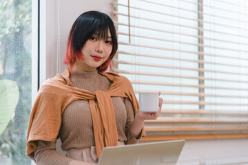 A beautiful Asian freelancer businesswoman is using a digital laptop near a window in her home office.