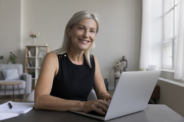 Positive confident mature freelance entrepreneur woman working at laptop, typing, sitting at home...