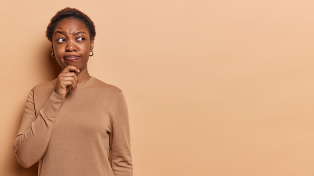 Doubtful dark skinned woman keeps hand on chin ponders difficult question tries to make right choice thinks about something wears casual jumper poses against brown background copy space for your text