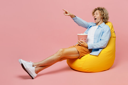 Full body young caucasian blond man wear blue shirt white t-shirt sit in bag chair point index finger aside on area eat pop corn watch movie film isolated on plain pastel light pink background studio.