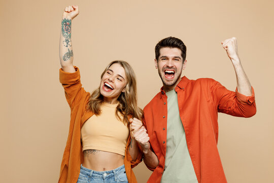 Young couple two friends family man woman wear casual clothes looking camera together do winner gesture celebrate clenching fists say yes isolated on pastel plain light beige color background studio.