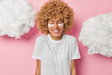 Positive woman with fair curly hair keeps eyes closed smiles toothily applies beauty patches under eyes to reduce wrinkles dressed in casual white t shirt isolated over pink background. Skin care