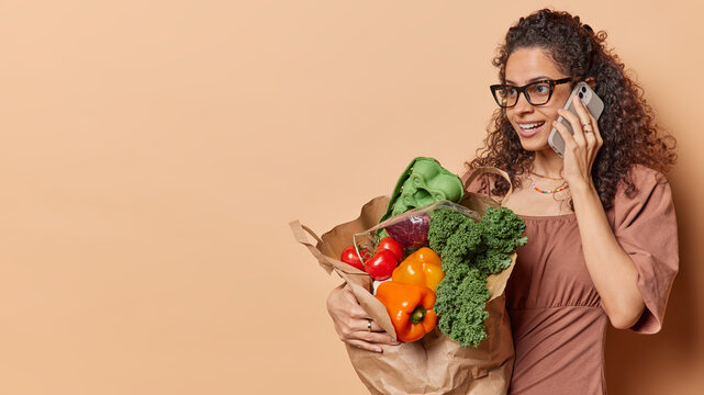 Glad Brazilian woman with cutly hair has telephone conversation carries grocery bag full of organic products wears spectacles and dress isolated over brown background copy space for your promotion