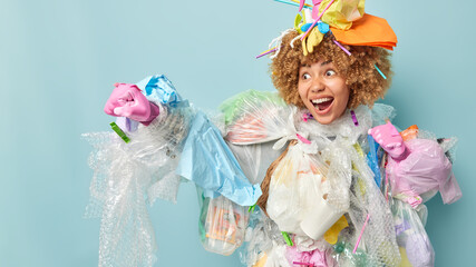 Crazy emotional curly haired woman makes fist pump exclaims loudly covered with plastic garbage tries to save environment from plastic pollution isolated over blue background. Ecology and recycling