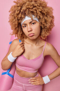 Discontent sportswoman dressed in tracksuut stands sweaty against pink background keeps hand on waist carries rolled karemat on shoulder focused unhappily at camera isolated. People and sport concept