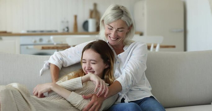 Cheerful loving grandma hugging teen granddaughter kid resting on sofa, tickling girl, embracing child, looking at camera, away, smiling, laughing, having fun, enjoying family leisure time