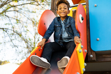 A cute dark-skinned boy between 5 and 6 years old with afro hair is sitting on a slide in an...