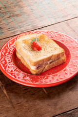 grilled ham and cheese sandwich on a red ceramic plate garnished with cherry tomatoes and oregano on a rustic wooden breakfast table at an angle