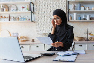 Obraz na płótnie Canvas Worried young woman in hijab checking her accounts, budget, expenses at home. Sitting at home in the kitchen with a laptop and documents. He holds his head in frustration.
